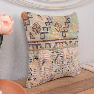 May include: A square cushion with a beige, green, and blue patterned design. The cushion has a textured appearance and is placed on a wooden surface. The cushion is part of a home decor collection.