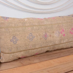 Op de afbeelding: Rechthoekig beige kussen met een patroon. Het kussen heeft een centrale band met herhaalde roze en blauwe bloemmotieven. Het ligt op een houten oppervlak, met een bloemstuk en een gevlochten mand zichtbaar.