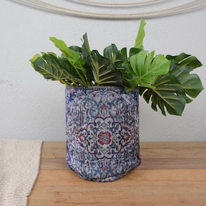 Peut inclure: Un pot cylindrique avec un motif bleu, rouge et blanc, rempli de feuilles vertes vibrantes. Le pot est posé sur une surface en bois, avec un textile de couleur neutre sur la gauche.