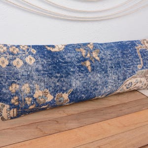 May include: A long, rectangular cushion with a distressed, vintage appearance. The cushion has a deep blue background with faded beige and gold floral patterns. It rests on a wooden surface, highlighting its decorative design.