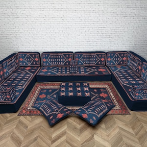 Puede incluir: Un sofá seccional azul marino con un diseño estampado en rojo y naranja. El conjunto incluye una alfombra, un otomán y dos cojines. El sofá está dispuesto en forma de U, adecuado para una sala de estar.