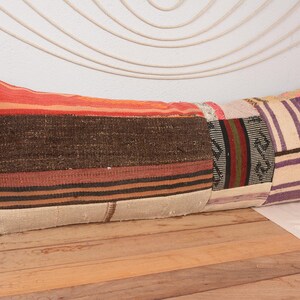 May include: A long, rectangular cushion with a patchwork design. The cushion features a variety of colours, including orange, brown, beige, and purple. The cushion is made of a woven fabric and has a rustic, bohemian style.