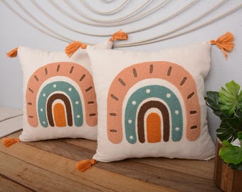Pode incluir: Duas almofadas brancas com desenhos de arco-íris bordados. Os arco-íris são em tons de laranja, marrom, turquesa e branco. As almofadas têm borlas laranja.