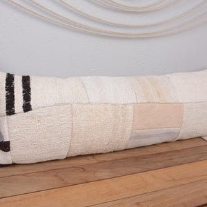 Puede incluir: Un cojín rectangular color crema con un diseño de patchwork. El cojín presenta una mezcla de texturas y tonos crema, beige y marrón. Dos rayas negras verticales están en un extremo. Está sobre una superficie de madera.