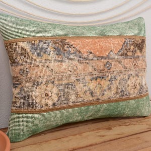 May include: Rectangular decorative pillow with a vintage rug design. The pillow features a central section with a faded orange and blue pattern, framed by green fabric. The pillow is placed on a wooden surface.