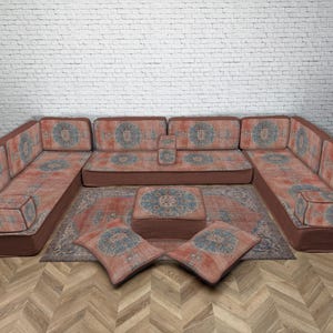 Puede incluir: Sofá seccional en forma de U con otomana y cojines a juego. Los muebles presentan una tela estampada en naranja rojizo y azul. El sofá y la otomana tienen una base marrón oscuro. Una alfombra estampada se coloca en el centro.