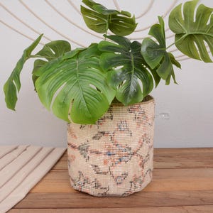 May include: A woven basket planter with a colorful geometric pattern holds a large green plant with broad, split leaves. The basket is beige with red, blue, and brown accents. The plant is a vibrant green.