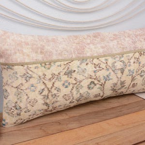 May include: A rectangular cushion with a vintage rug design. The top section is a light pink, while the bottom features a cream-coloured pattern with blue and brown accents. A beige border separates the two sections. The cushion is placed on a wooden surface.