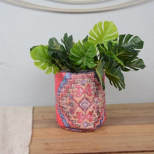 May include: A cylindrical plant pot with a colorful, patterned design, holding vibrant green leafy plants. The pot is made of fabric and sits on a wooden surface. The pot's design features red, blue, and beige hues.