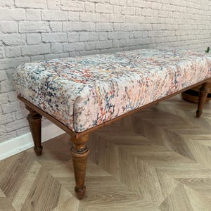 May include: A wooden bench with turned legs and a floral patterned fabric seat. The bench is sitting on a wood floor with a herringbone pattern.