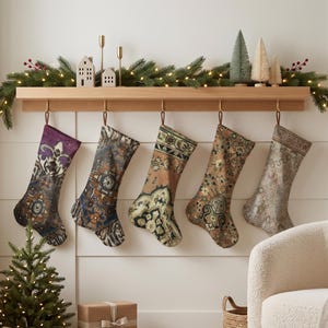 Puede incluir: Cinco calcetines navideños colgados de una repisa de madera, cada uno con un patrón de alfombra estilo vintage distinto, intrincado y multicolor. Los calcetines muestran paletas de colores variadas que incluyen tonos morados, azul oscuro, naranja quemado y beige.