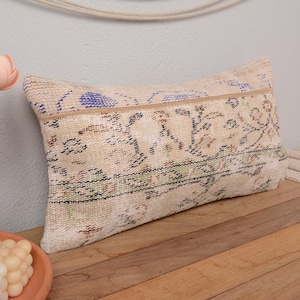 Puede incluir: Una almohada rectangular con fondo crema y un patrón floral en tonos azules, verdes y marrones. La almohada está hecha de un tejido tejido y tiene un borde con flecos.