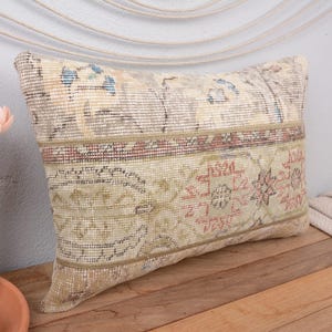 May include: Rectangular decorative pillow with a vintage rug design. The pillow features a beige background with floral and geometric patterns in shades of red, blue, and green. The pillow is placed on a wooden surface.