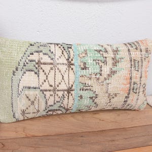 Może przedstawiać: Prostokątna poduszka dekoracyjna o wyglądzie vintage i postarzanym. Poduszka ma wzór patchwork z wyblakłymi kolorami, w tym jasną zielenią, beżem i odcieniami pomarańczu i błękitu. Wzór zawiera elementy geometryczne i kwiatowe.