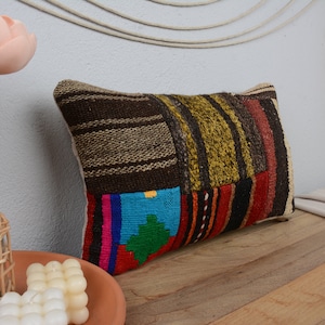 May include: Rectangular decorative pillow with a patchwork design. The pillow features a mix of brown, green, red, and blue fabric sections. The pillow is placed on a wooden surface, with decorative elements in the background.