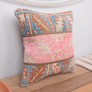 May include: A square decorative pillow with a vintage rug design. The pillow features a central pink panel, bordered by sections with blue, brown, and beige geometric patterns. The pillow is placed on a wooden surface.