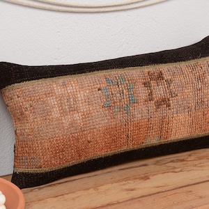 Puede incluir: Cojín decorativo rectangular con un diseño tejido y texturizado. El cojín presenta un panel central en tonos naranja y marrón, con un borde marrón oscuro. El diseño incluye formas geométricas en azul y marrón.