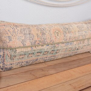 May include: A long, rectangular pillow with a faded, patterned design in shades of peach, blue, and green. The pillow has a textured appearance and is accented with a thin, light-colored band. It rests on a wooden surface.