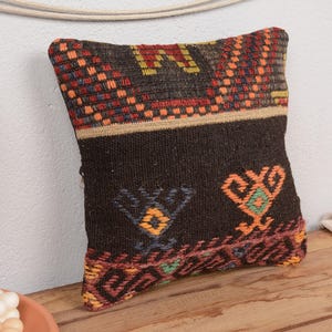 May include: A decorative, rectangular pillow with a dark brown background and colorful geometric patterns. The pillow features orange, red, yellow, and blue accents. The pillow is placed on a wooden surface.