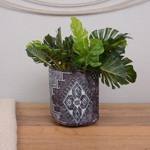 Pode incluir: Um vaso decorativo para plantas com um padrão geométrico em tons de cinza e branco, preenchido com folhas verdes grandes. O vaso está sobre uma superfície de madeira, com um cobertor e uma parede de cor neutra no fundo.