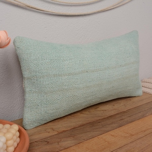 May include: A light green throw pillow with a woven texture. The pillow is on a wooden surface.