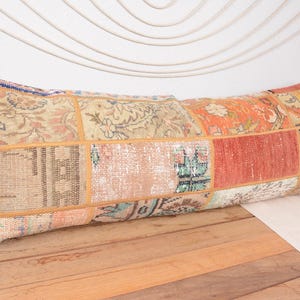 May include: Rectangular patchwork pillow with a variety of colors including orange, beige, and red. The pillow has a vintage, distressed look with visible stitching and a mix of patterned fabric squares. It rests on a wooden surface.