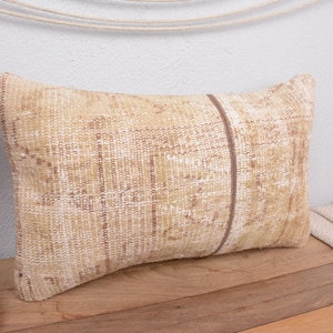 May include: Rectangular beige pillow with a brown stripe down the center. The pillow has a woven texture and is placed on a wooden surface. Decorative flowers and a woven basket are visible in the background.