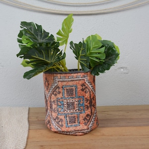 Puede incluir: Una maceta cilíndrica con un diseño naranja y azul estampado, que contiene una planta de hojas verdes. La maceta está sobre una superficie de madera, con un textil de color neutro a la izquierda.