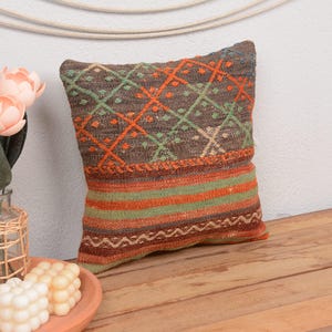 Puede incluir: Un cojín decorativo cuadrado con un patrón geométrico en tonos marrón, naranja y verde. El cojín está colocado sobre una superficie de madera, con un jarrón de flores rosas y velas en primer plano. El diseño del cojín presenta rombos y rayas.