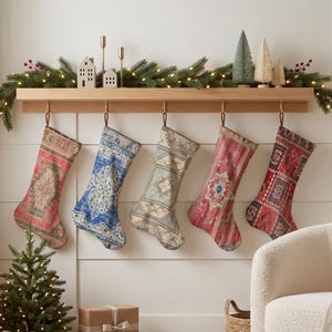 Puede incluir: Cinco calcetines navideños colgados de una repisa de madera, cada uno con un patrón único, intrincado y multicolor que recuerda a las alfombras tejidas. Los calcetines muestran varios tonos de rojo, azul, beige y crema en diseños geométricos.