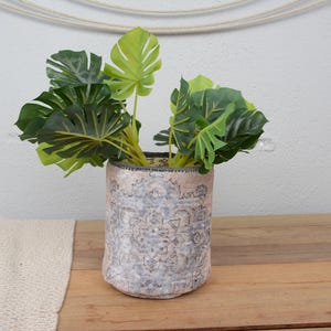 May include: A decorative cylindrical planter with a distressed, patterned exterior, holding vibrant green artificial Monstera leaves. The planter sits on a wooden surface, adding a touch of nature-inspired decor to any space.