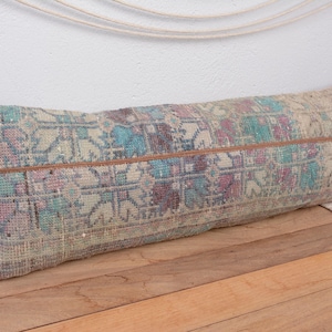 Op de afbeelding: Langwerpig decoratief kussen met een vintage patroon in blauw, paars en beige. Het kussen heeft een bruine, leren streep in het midden. Het kussen ligt op een houten oppervlak.
