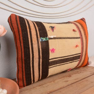 May include: Rectangular cushion with a woven design. The cushion features a cream-coloured centre with dark brown and tan stripes. The edges are accented with orange and brown stripes. Small colourful accents are scattered across the surface.