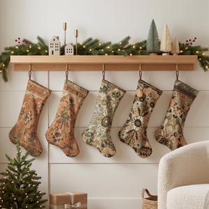 May include: Five Christmas stockings with intricate, vintage-inspired floral and geometric patterns in muted tones of rust, sage green, cream, and blue, hanging from a light wood mantel shelf decorated with a lit pine garland and small ornaments.
