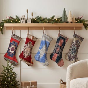 Op de afbeelding: Vijf kerstsokken hangen aan een houten schoorsteenmantel, elk met een opvallend geweven tapijtpatroon in rood, blauw, crème en donkere tinten. De sokken hangen aan leren lussen aan koperen haken aan een plank van lichtgekleurd hout.