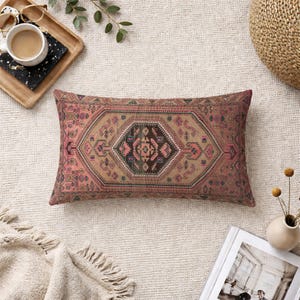 Könnte beinhalten: Rechteckiges Zierkissen mit Vintage-Teppichdesign. Das Kissen hat ein zentrales geometrisches Muster in Braun-, Rosa- und Schwarztönen mit aufwendigen Bordürendetails. Das Kissen liegt auf einem neutralfarbenen Teppich.