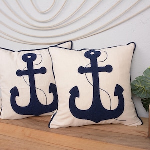 May include: Two white cushion covers with navy blue embroidered anchor designs. The cushions are on a wooden surface.