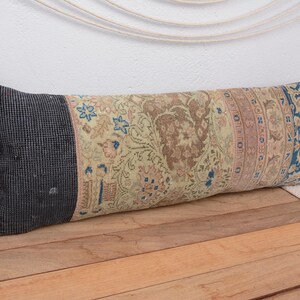 May include: A long, rectangular pillow with a patterned design. The pillow features a dark gray end, a central section with a beige and brown floral pattern, and a blue and beige patterned end. The pillow is on a wooden surface.