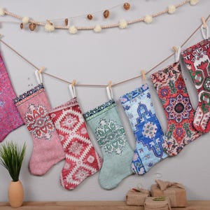 Peut inclure: Six chaussettes de Noël colorées avec des motifs complexes sont suspendues à une guirlande de pompons blancs. Les chaussettes sont en tissu avec une variété de motifs, y compris des motifs floraux, géométriques et paisley.
