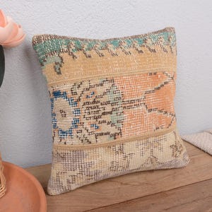 Op de afbeelding: Een vierkant decoratief kussen met een patchwork ontwerp. Het kussen heeft een mix van kleuren, waaronder beige, oranje, blauw en groen. Het kussen ligt op een houten oppervlak, met een vaas met bloemen op de achtergrond.