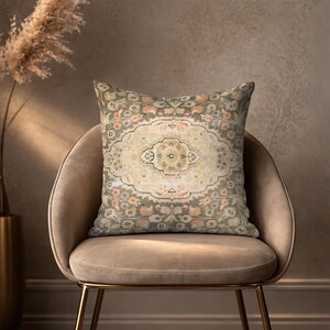 Könnte beinhalten: Ein dekoratives Zierkissen mit einem Vintage-Teppichdesign. Das Kissen hat ein zentrales Medaillon in Beige und Creme, umgeben von einem Rand aus gedämpften Grüntönen, Orange und floralen Mustern. Es liegt auf einem beigen Samtstuhl mit goldenen Beinen.