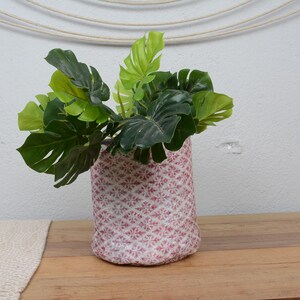 Könnte beinhalten: Eine zylindrische Vase mit einem rosa-weißen Muster enthält lebendige grüne tropische Blätter. Die Vase steht auf einer Holzoberfläche und schafft eine natürliche und dekorative Präsentation. Die Blätter haben eine Mischung aus dunklen und hellgrünen Farben.