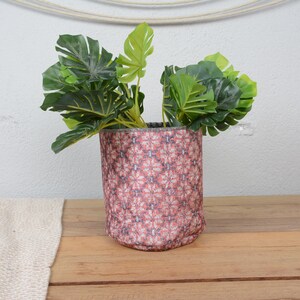 May include: A cylindrical plant pot with a floral pattern in shades of pink and gray, holding a vibrant green plant with large leaves. The pot sits on a wooden surface.
