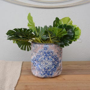 May include: A round planter with a blue and pink patterned design, filled with vibrant green tropical leaves. The planter sits on a wooden surface, offering a touch of nature-inspired decor.