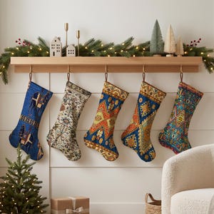 Può includere: Cinque calze di Natale appese a una mensola in legno, ognuna caratterizzata da distinti motivi intrecciati. I design includono blu con motivi di cammelli, crema con dettagli floreali e tre varianti di stampe geometriche multicolori.