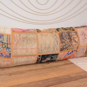 Puede incluir: Cojín rectangular de patchwork con una variedad de colores y patrones. El cojín presenta una mezcla de tonos beige, azul, naranja y rosa, con un aspecto texturizado. Se coloca sobre una superficie de madera.