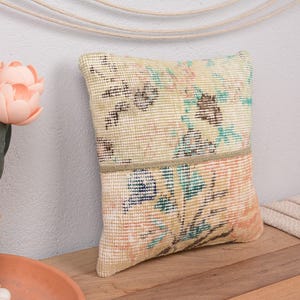 May include: A decorative pillow with a floral pattern in shades of beige, peach, and teal. The pillow is rectangular and appears to be made of woven fabric. The pillow is on a wooden surface.