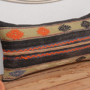 May include: Rectangular decorative pillow with a woven design. The pillow features a black background with orange, green, and blue geometric patterns. The pillow is placed on a wooden surface.