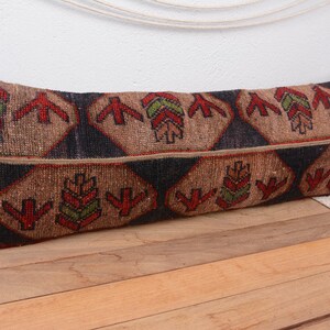 May include: A rectangular, patterned pillow with a brown background and geometric designs in red, green, and black. The pillow is placed on a wooden surface.