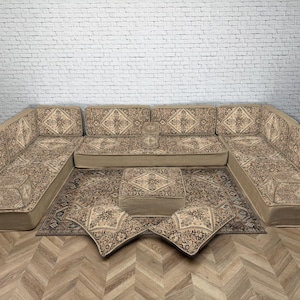 以下が含まれることがあります： 模様入りのクッションとマッチするラグが付いたベージュのセクショナルソファ。ソファはロープロファイルで、U字型に配置されています。ラグとクッションは、繰り返しの幾何学模様が特徴です。